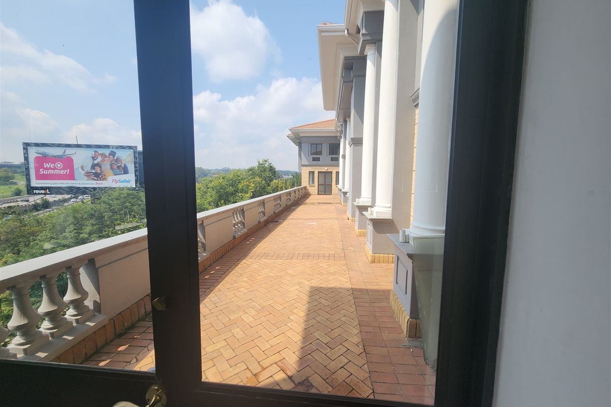A spacious outdoor balcony featuring herringbone brick paving, classical columns, and a balustrade, offering an elevated view of the surrounding area.