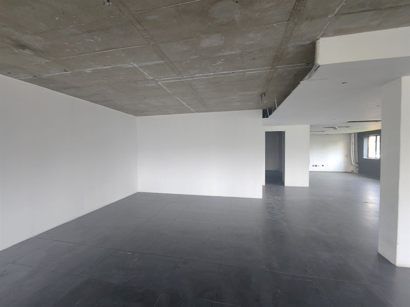 An open-plan office space featuring an exposed concrete ceiling, large format dark floor tiles, and a neutral white wall finish.