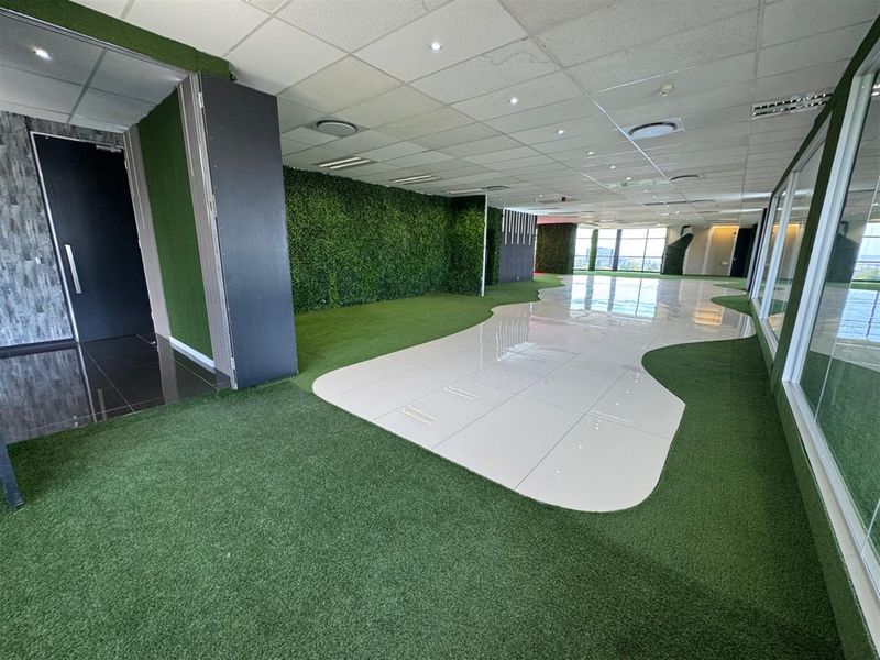 Spacious open-plan office area featuring a unique combination of artificial grass and ceramic tile flooring, glass partitions, and integrated green wall accents.