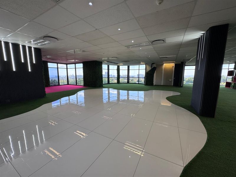 Spacious open-plan office area featuring floor-to-ceiling windows with panoramic views, modern tiled and artificial turf flooring, and contemporary vertical wall accents.