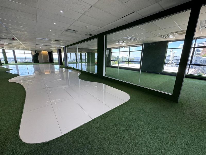 Spacious open-plan office area featuring a modern mix of tiled and artificial turf flooring, glass partitions, and large windows with natural light.