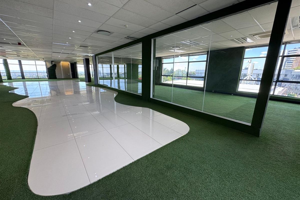 Spacious open-plan office area featuring a modern mix of tiled and artificial turf flooring, glass partitions, and large windows with natural light.