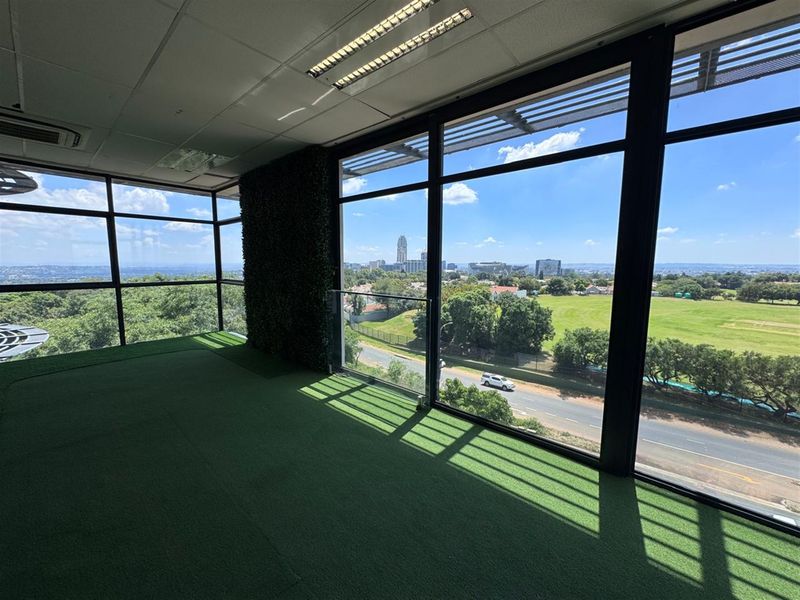 Bright office space featuring floor-to-ceiling windows with expansive city views, artificial grass flooring, and a decorative green wall feature.