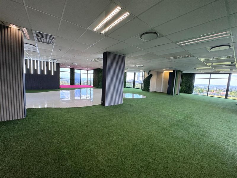 Spacious open-plan office area featuring floor-to-ceiling windows with panoramic views, modern wall paneling, vertical garden accents, and a mix of artificial grass and tiled flooring.