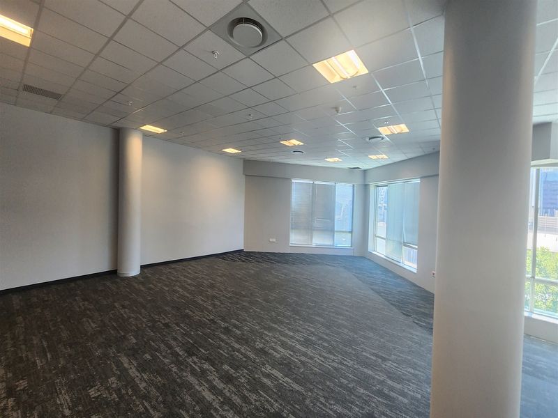 A spacious, open-plan office area featuring a suspended ceiling, recessed lighting, carpeted flooring, and large windows providing ample natural light.
