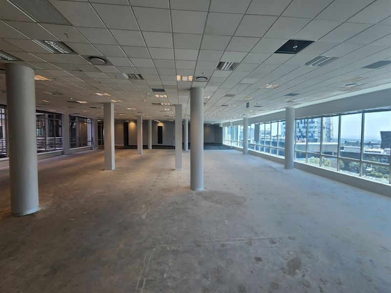 A large, open-plan office space featuring structural columns, a suspended ceiling with recessed lighting, and expansive windows offering natural light and city views.