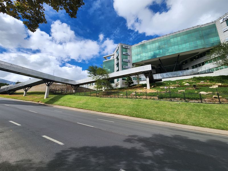 Modern office building exterior featuring a glass facade and a prominent pedestrian skybridge, set above a well-maintained landscaped embankment and road frontage.