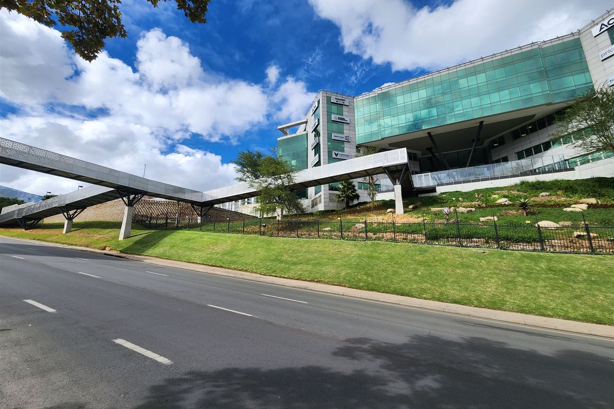 Modern office building exterior featuring a glass facade and a prominent pedestrian skybridge, set above a well-maintained landscaped embankment and road frontage.