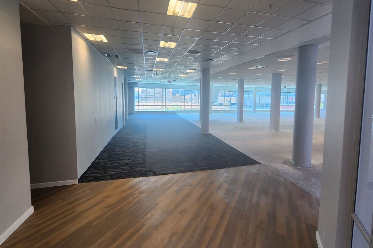 Spacious open-plan office area featuring large windows, structural columns, a suspended ceiling, and a mix of carpet and wood-look flooring.