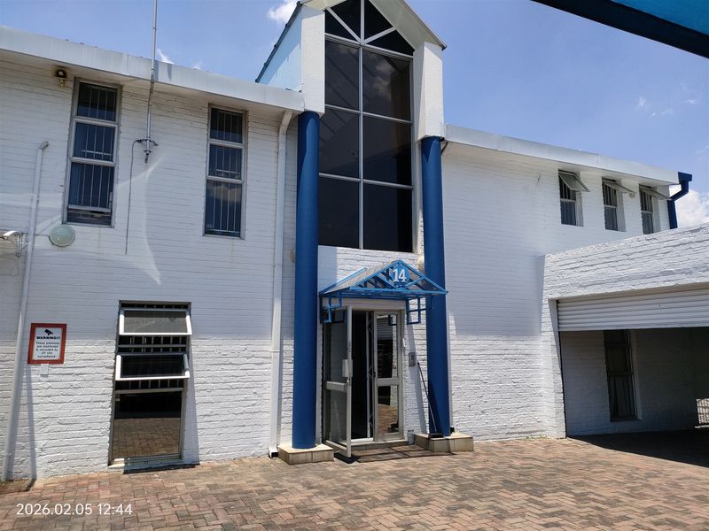 Front elevation of an industrial property featuring a paved entrance, glass facade, security bars, and a roller shutter door for vehicle access.