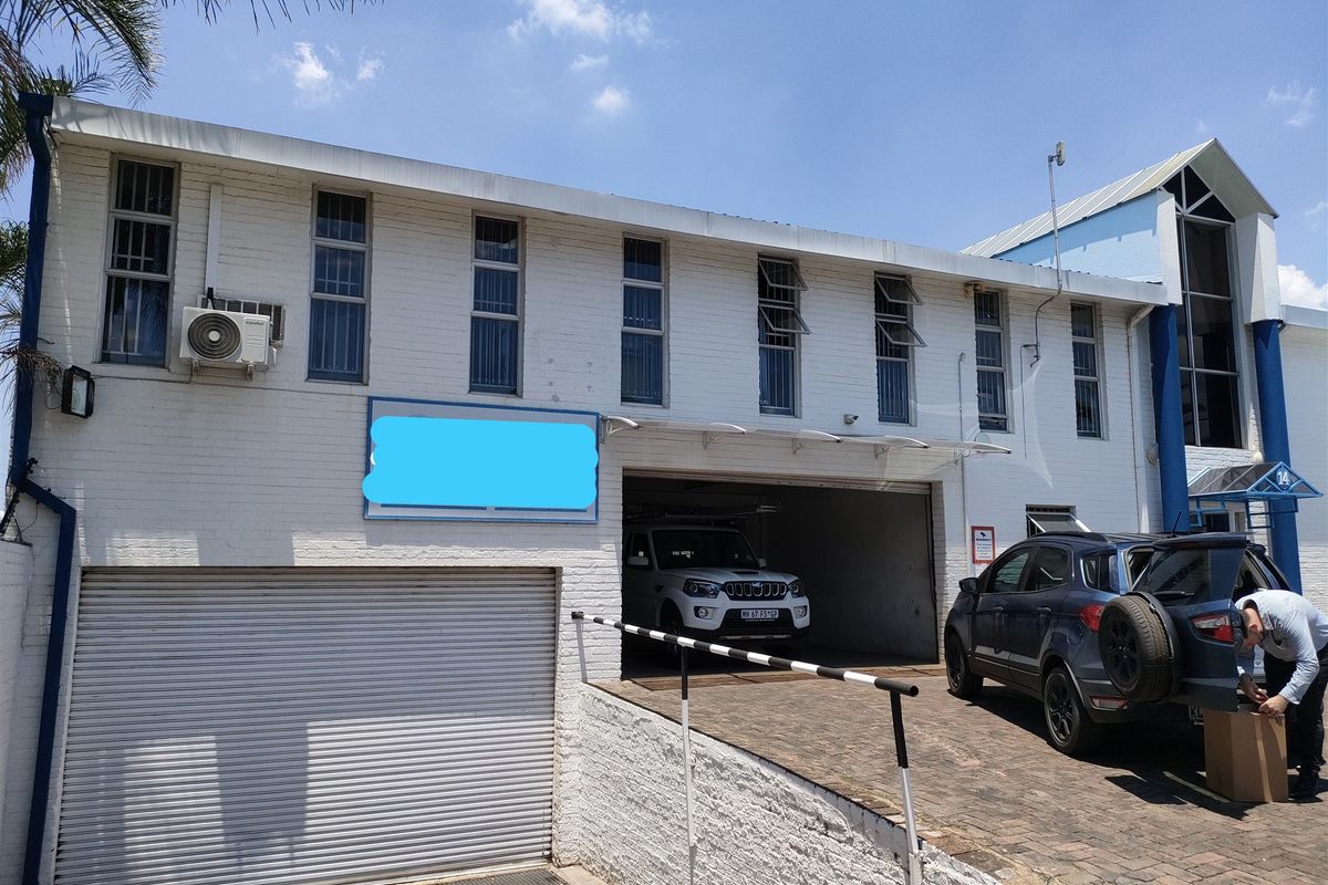 Front elevation of an industrial property featuring a roller shutter door, paved driveway access, and white painted brick facade with multiple windows.