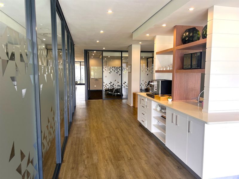 Modern office corridor featuring glass partition walls, wood-look flooring, recessed lighting, and a sleek built-in kitchenette area with stone countertops.