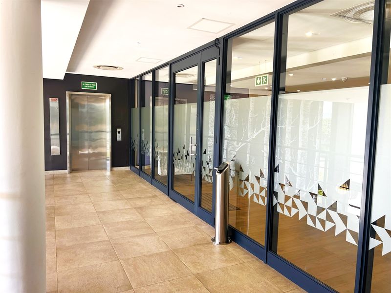 Modern office hallway featuring tiled flooring, a stainless steel elevator, and floor-to-ceiling glass partitions with decorative frosted film.