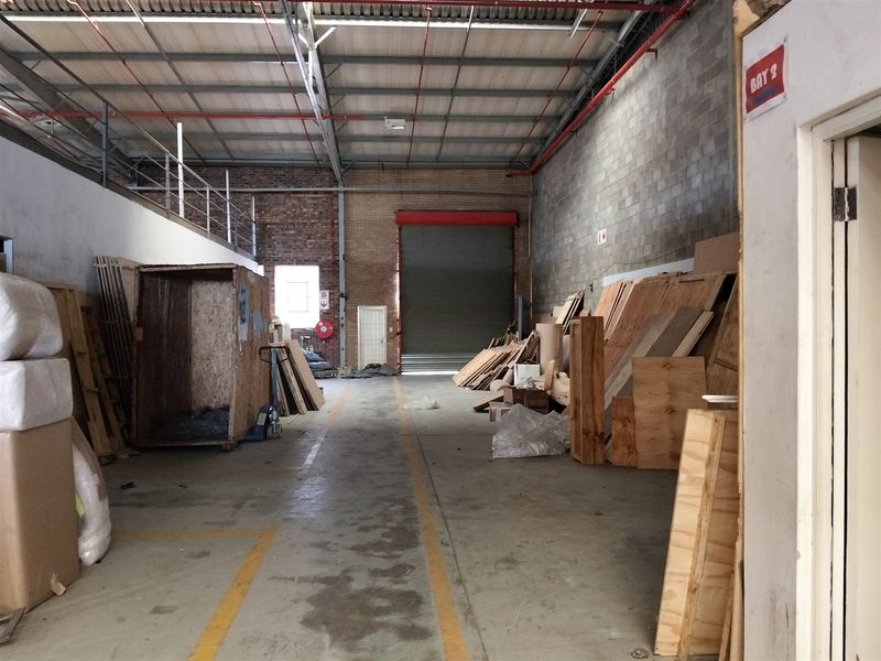 Spacious industrial warehouse unit featuring high ceilings with exposed steel trusses, a large roller shutter door, and a mezzanine level for additional storage.
