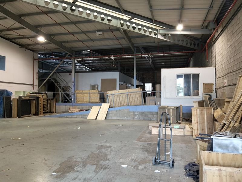 Spacious industrial warehouse area featuring high ceilings with exposed steel trusses, concrete flooring, a mezzanine level, and integrated fire sprinkler systems.