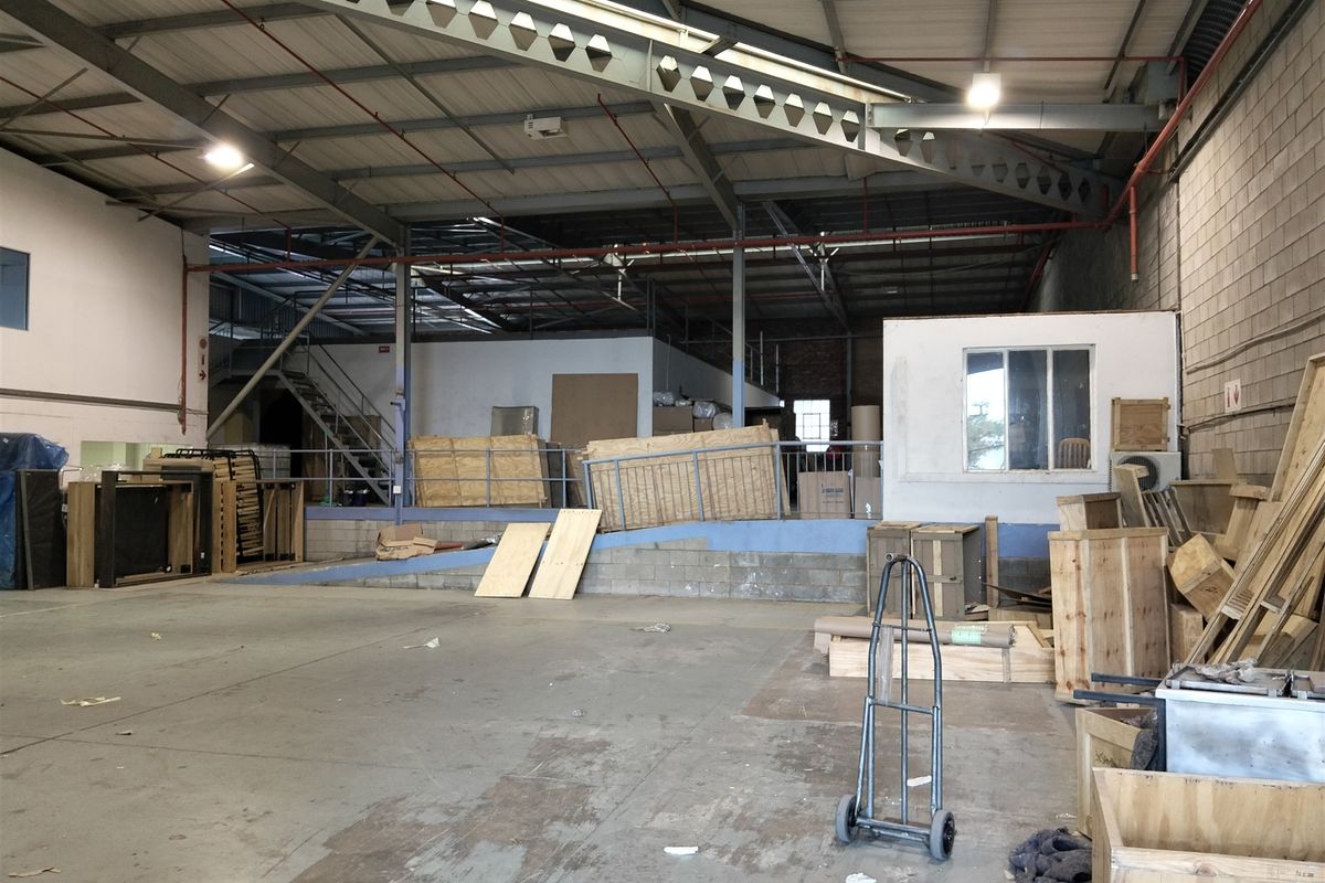 Spacious industrial warehouse area featuring high ceilings with exposed steel trusses, concrete flooring, a mezzanine level, and integrated fire sprinkler systems.