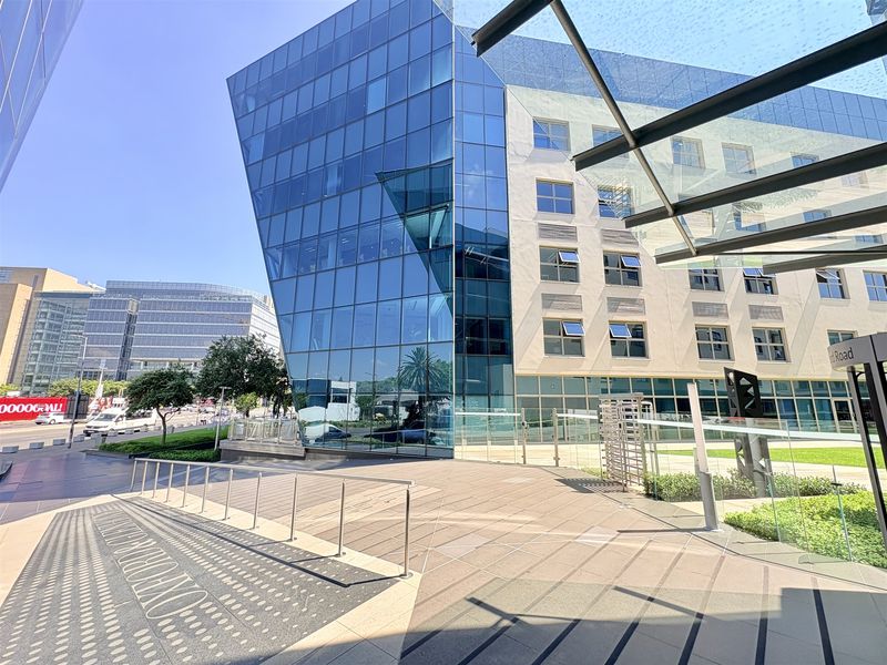 Modern office building exterior featuring a striking glass curtain wall facade, paved pedestrian plaza, and a contemporary glass entrance canopy.
