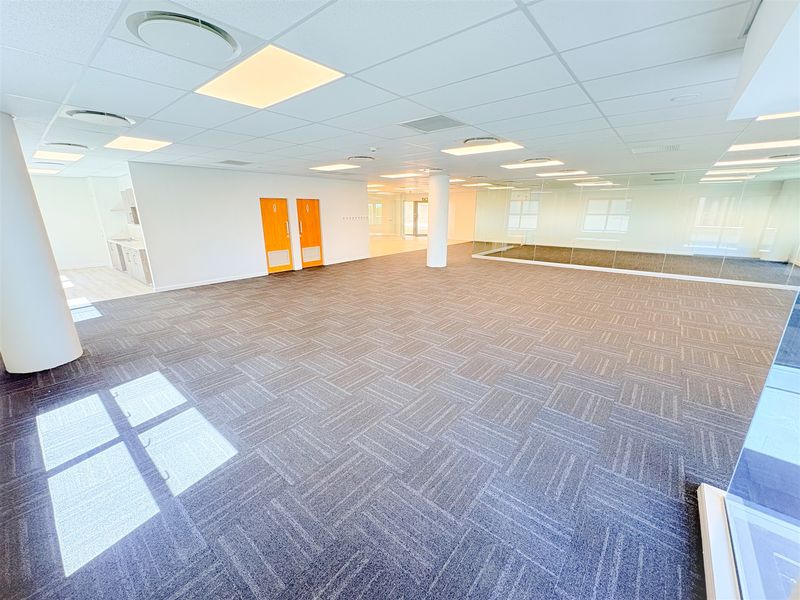 Spacious open-plan office area featuring carpet tiles, recessed lighting, a glass partition wall, and a small integrated kitchenette.