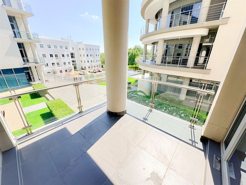 A spacious, modern balcony featuring tiled flooring, a sleek glass and stainless steel balustrade, and views over the office park grounds.