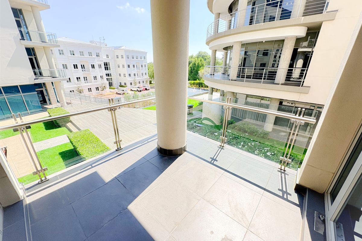 A spacious, modern balcony featuring tiled flooring, a sleek glass and stainless steel balustrade, and views over the office park grounds.