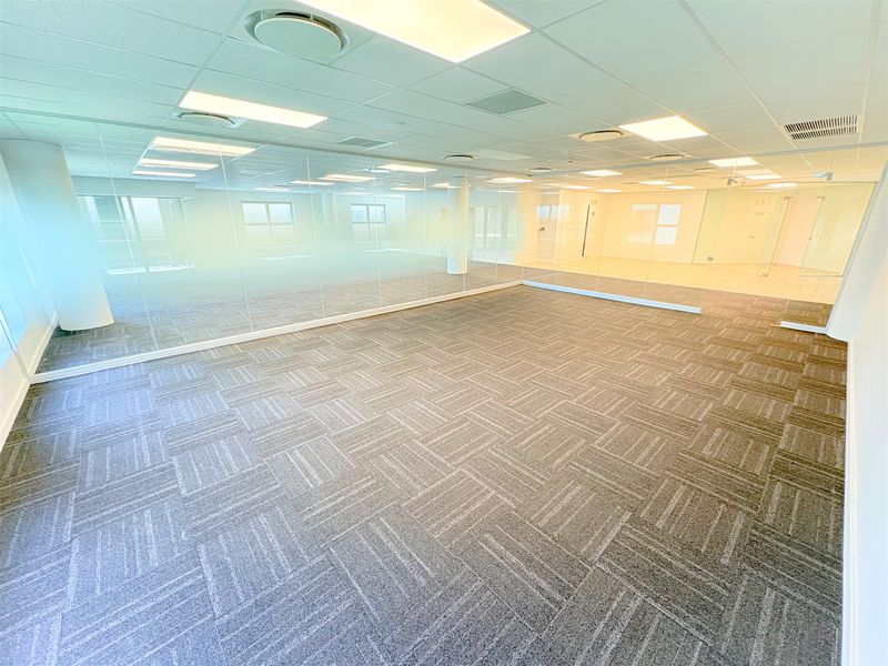 A spacious, empty office area featuring modern glass partitioning, carpet tile flooring, and a suspended ceiling with integrated lighting and air conditioning.