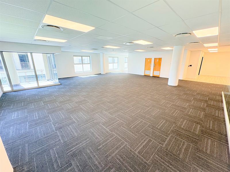 A spacious, open-plan office area featuring neutral carpet tiles, a suspended ceiling with recessed lighting, and large windows providing ample natural light.