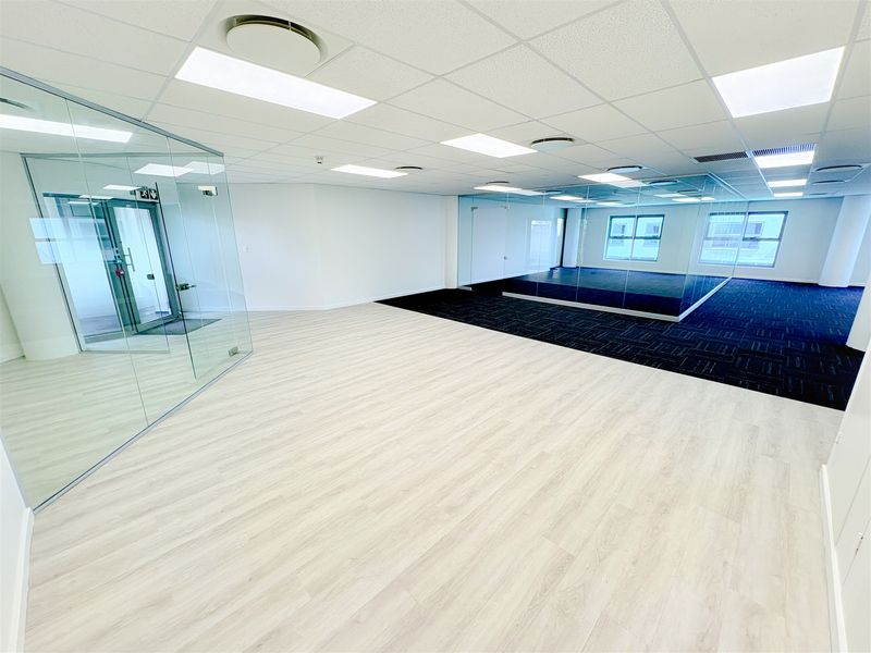 Modern open-plan office space featuring glass partitioning, a mix of laminate and carpet flooring, and recessed lighting in a suspended ceiling.