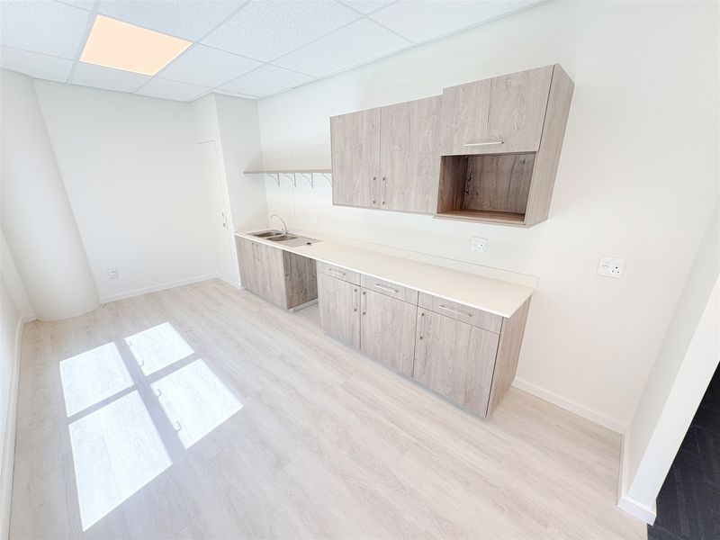 A clean, modern office kitchenette featuring light wood-grain cabinetry, a stainless steel sink, and durable laminate flooring in a bright, open workspace.