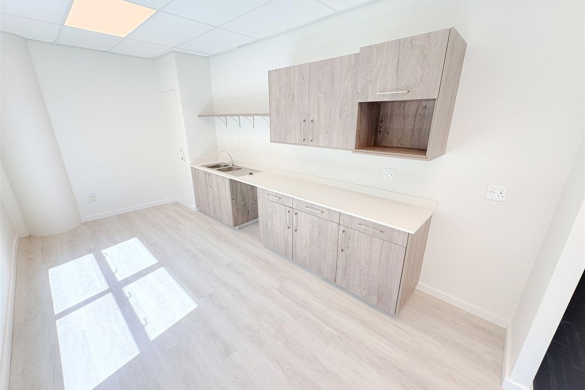 A clean, modern office kitchenette featuring light wood-grain cabinetry, a stainless steel sink, and durable laminate flooring in a bright, open workspace.