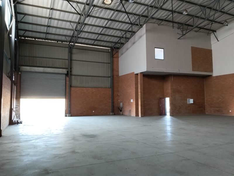 Spacious industrial warehouse unit featuring high ceilings with exposed steel trusses, concrete flooring, exposed brick walls, and a large roller shutter door.