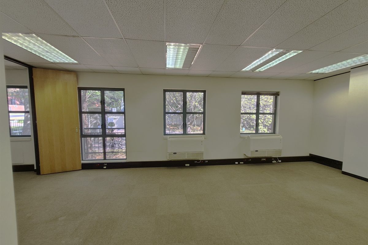 A spacious, empty office area featuring a suspended ceiling, recessed lighting, carpeted flooring, and under-window HVAC units with natural light.