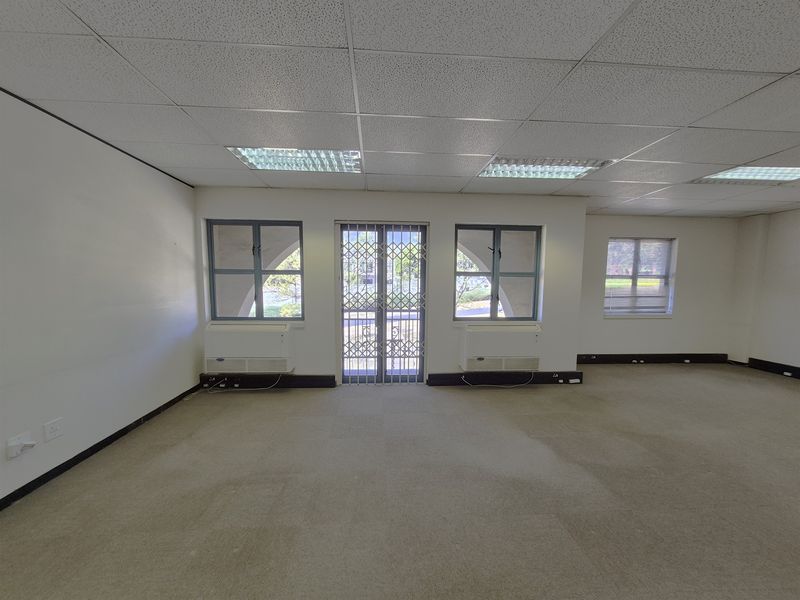 An empty, open-plan office space featuring a suspended ceiling, recessed lighting, carpeted flooring, and security-gated access to the exterior.