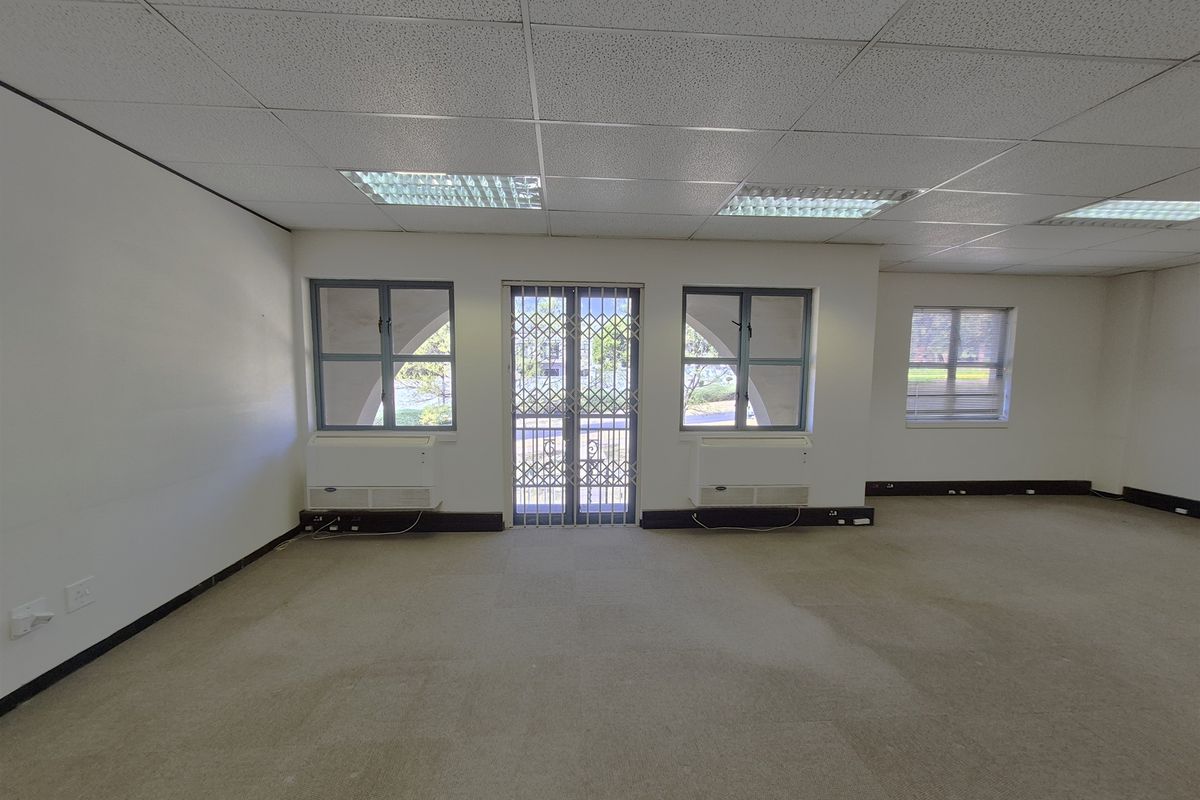 An empty, open-plan office space featuring a suspended ceiling, recessed lighting, carpeted flooring, and security-gated access to the exterior.
