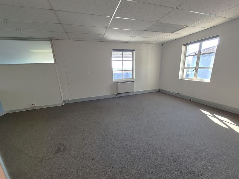 A vacant office space featuring a suspended ceiling, carpeted flooring, wall-mounted air conditioning, and natural light from multiple windows.