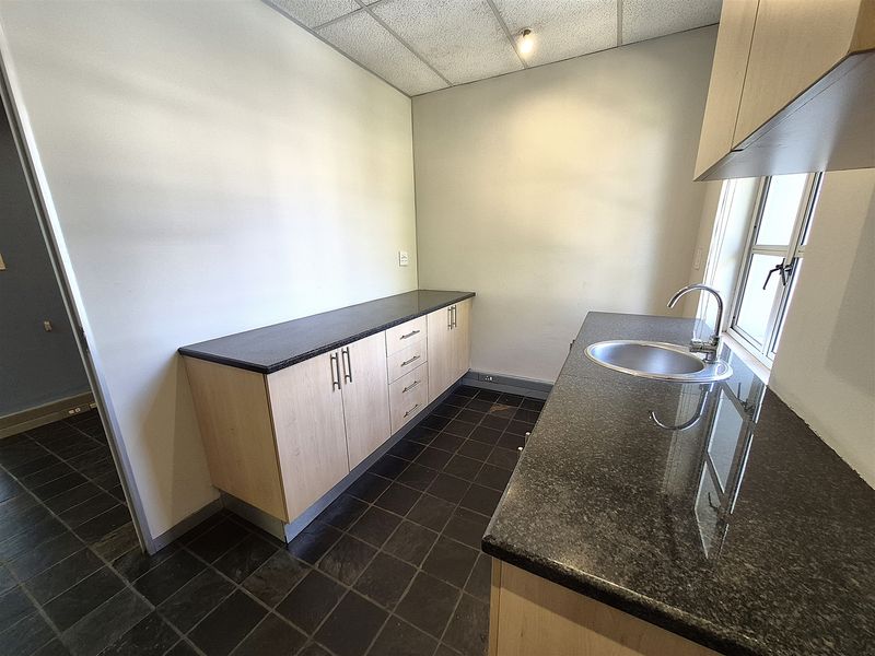 A clean, functional kitchenette featuring light wood cabinetry, dark granite countertops, a stainless steel sink, and durable tiled flooring.