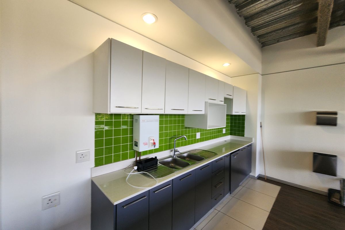 A clean, modern office kitchenette featuring white upper cabinets, dark lower cabinetry, a stainless steel double sink, and a vibrant green tiled splashback.