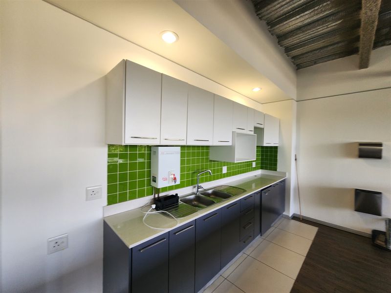 A clean, modern office kitchenette featuring white upper cabinets, dark lower cabinetry, a stainless steel double sink, and a vibrant green tiled splashback.