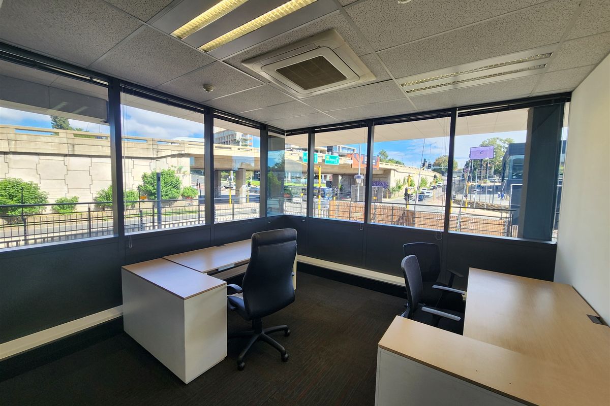 A bright corner office space featuring large windows with urban views, suspended ceilings, integrated air conditioning, and carpeted flooring.