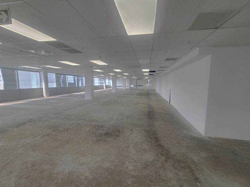 A large, open-plan office space featuring a suspended ceiling, recessed lighting, and expansive windows, currently in a shell condition with unfinished concrete flooring.