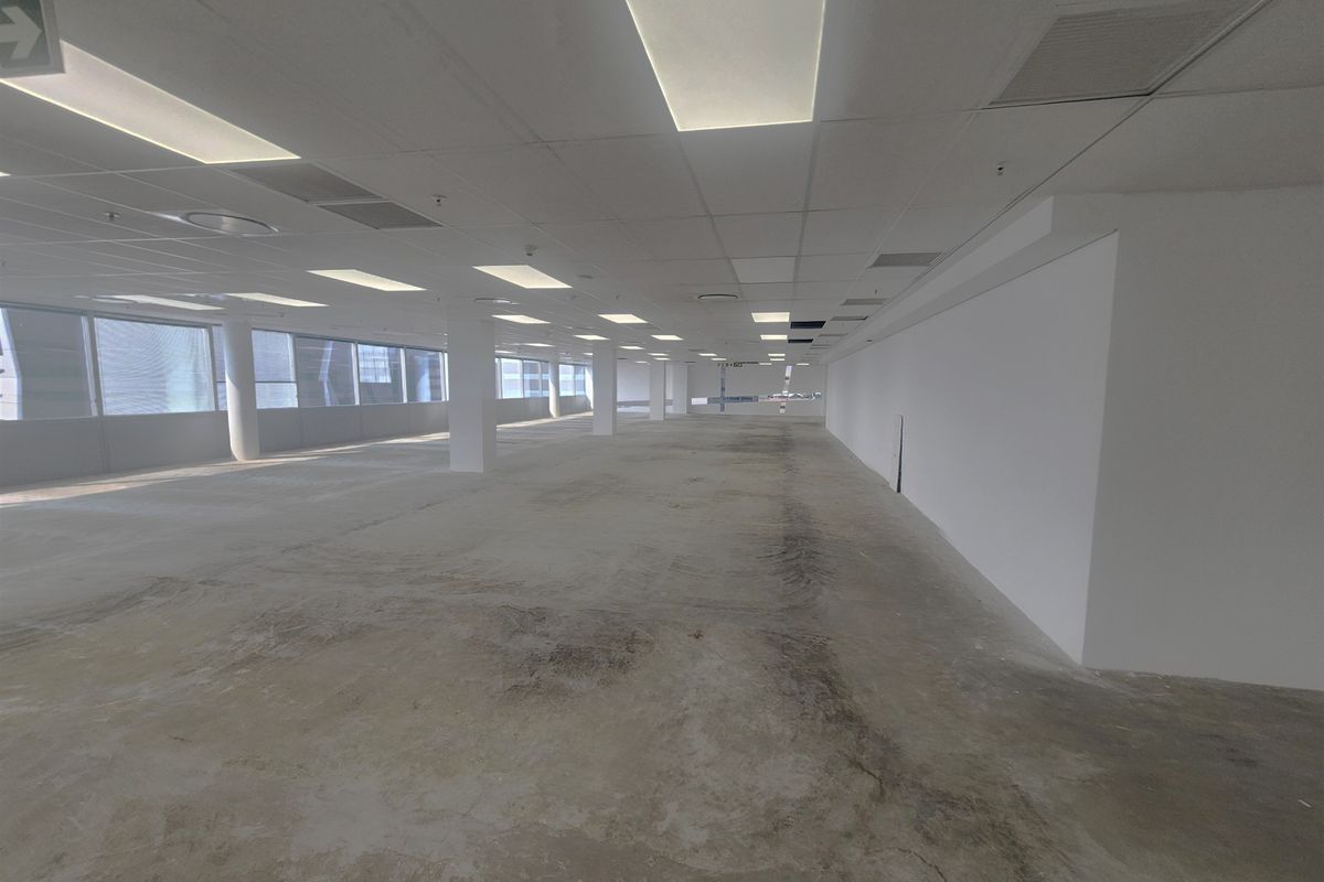 A large, open-plan office space featuring a suspended ceiling, recessed lighting, and expansive windows, currently in a shell condition with unfinished concrete flooring.