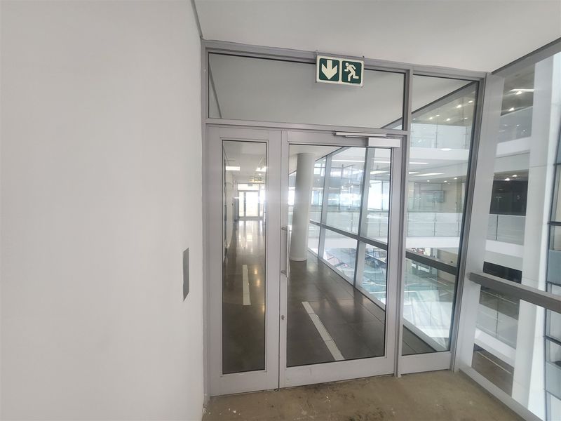 A modern office hallway featuring glass partitioning, aluminium framed doors, and clear views into the adjacent atrium space.