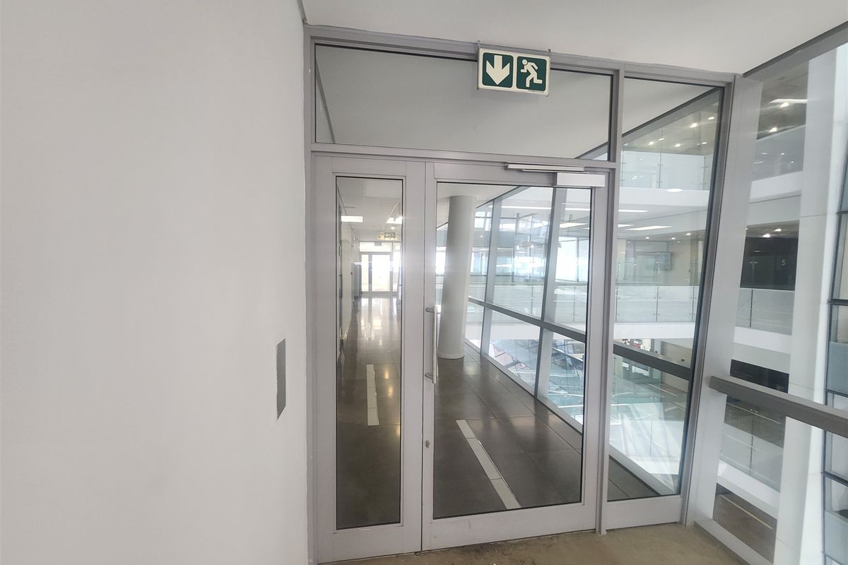 A modern office hallway featuring glass partitioning, aluminium framed doors, and clear views into the adjacent atrium space.
