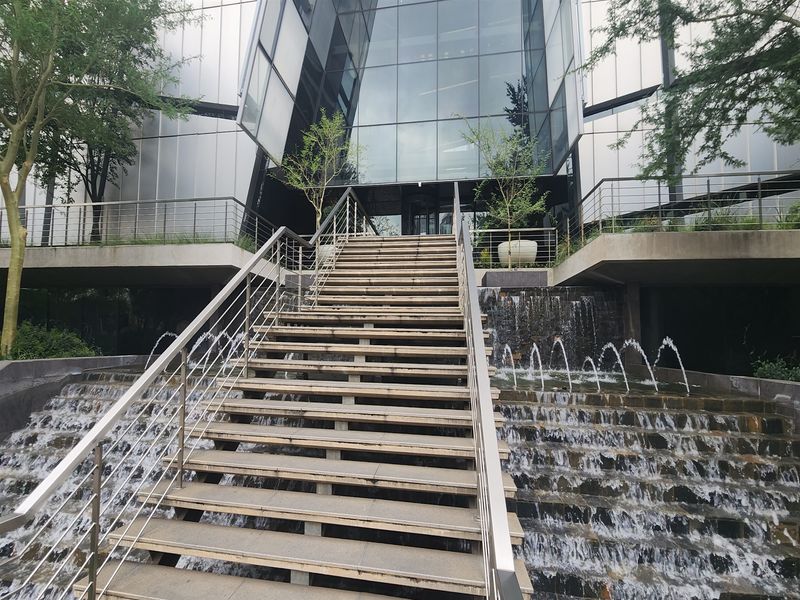 Modern office building entrance featuring a striking glass facade, tiered water feature, and a prominent central staircase with stainless steel railings.