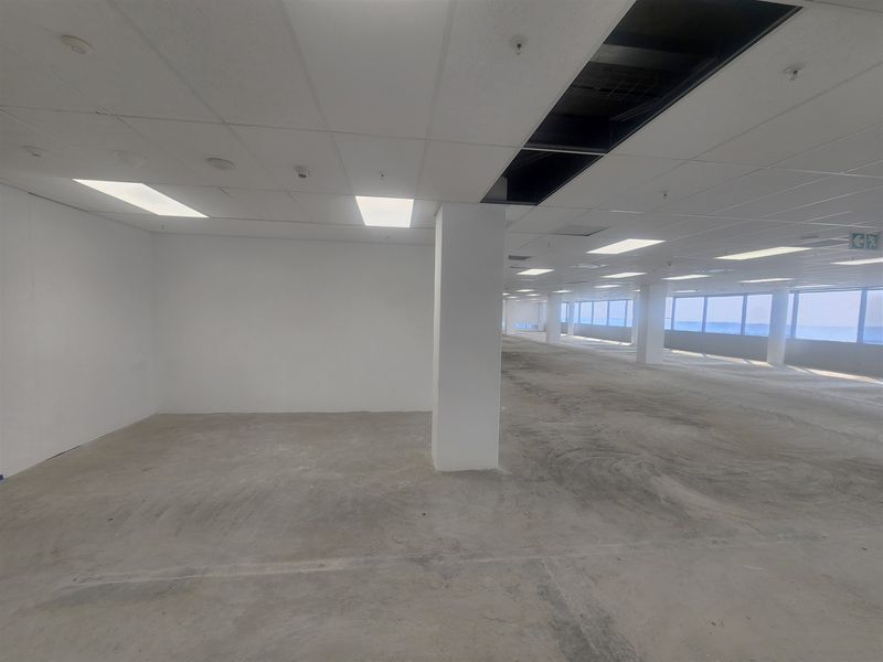 An expansive, open-plan office space featuring large windows, suspended ceilings, and unfinished concrete flooring, currently in a vacant shell condition.