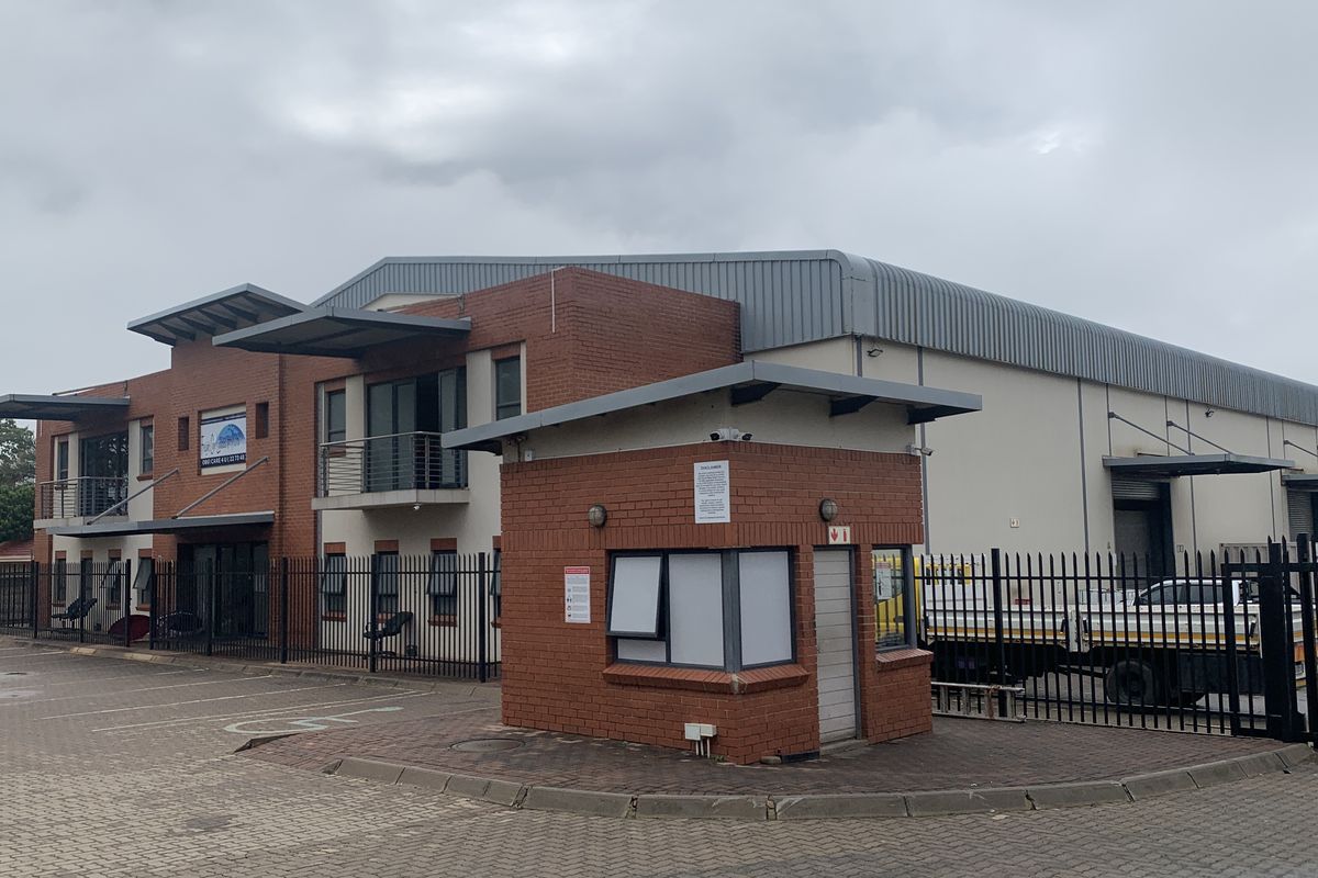 Front view of an industrial property featuring a brick security guard house, paved parking area, perimeter fencing, and large warehouse facility with roller shutter doors.