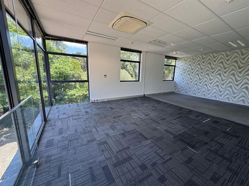 A bright, open-plan office space featuring carpet tiles, a suspended ceiling with integrated air conditioning, and large windows offering natural light and greenery views.
