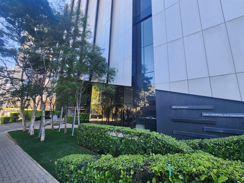 Modern office building exterior featuring a glass curtain wall, sleek cladding panels, and well-maintained landscaped hedging along the paved entrance walkway.