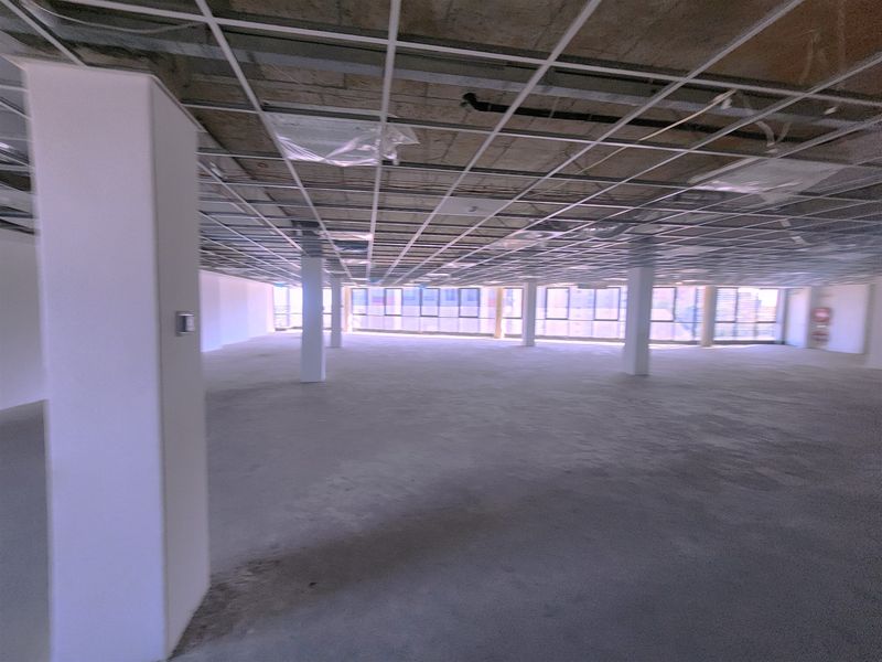 An expansive, open-plan office space in shell condition, featuring a suspended ceiling grid, structural columns, and large windows providing ample natural light.