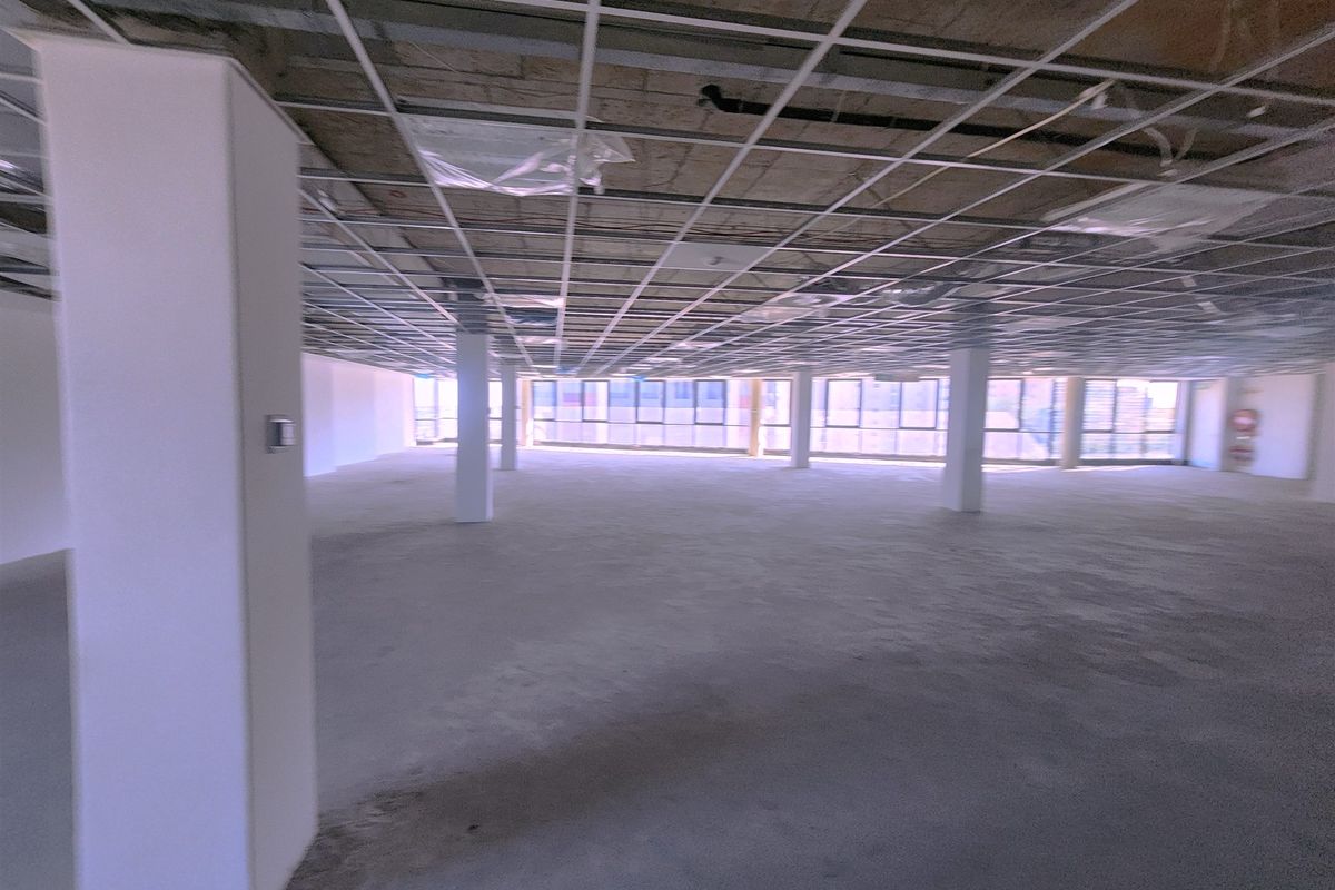 An expansive, open-plan office space in shell condition, featuring a suspended ceiling grid, structural columns, and large windows providing ample natural light.