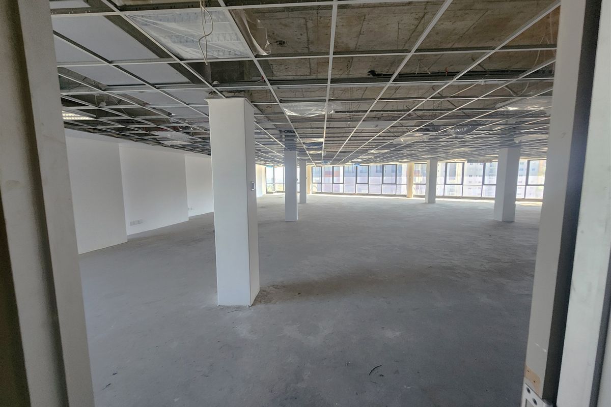 An expansive, open-plan office space currently in shell condition, featuring a suspended ceiling grid, structural columns, and large windows providing ample natural light.